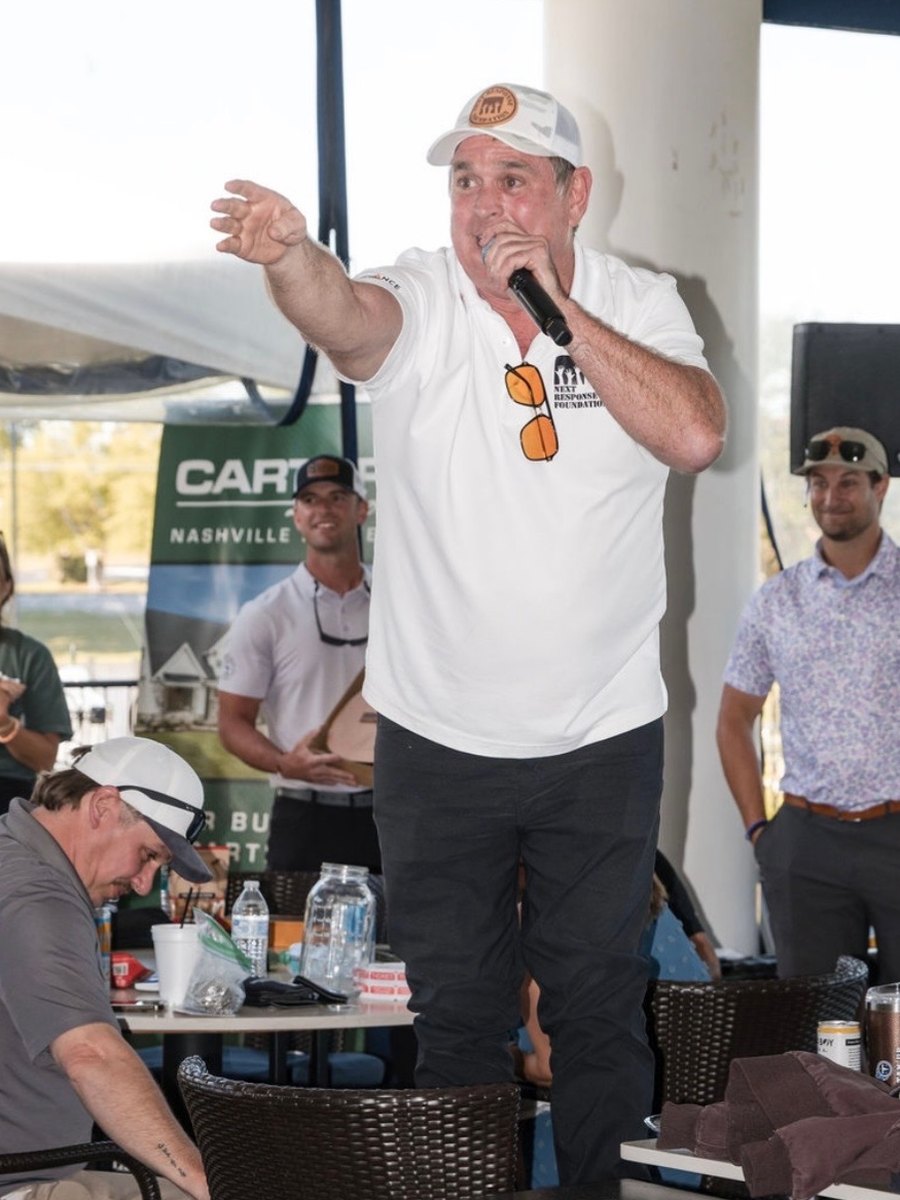 Todd Nichols calling bids from a clubhouse patio at a charity golf tournament, arm outstretched toward a bidder
