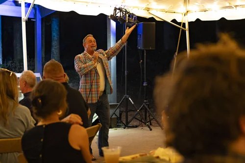 Todd Nichols under a tent at an outdoor benefit, holding up a live auction item to a seated crowd