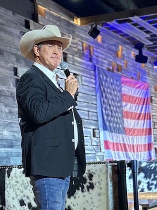 Todd Nichols on stage in a hat and dark jacket, microphone in hand, with an American flag on the wall behind him